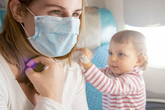Mujer con mascarilla sentada en un avión mientras un bebé intenta jugar con ella